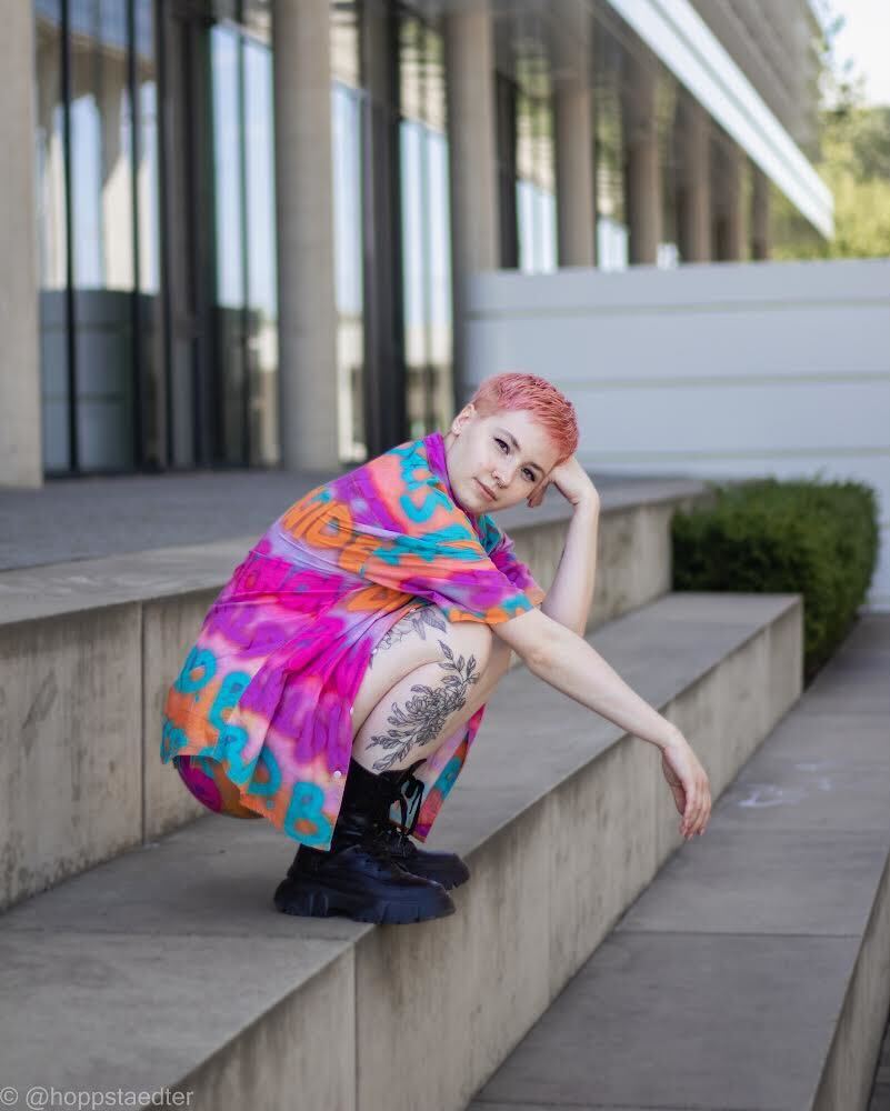 Porträt von Basja, der neuen Resident Tattoo-Künstlerin, in einem urbanen Umfeld mit sichtbarem Tattoo auf ihrem Oberschenkel.