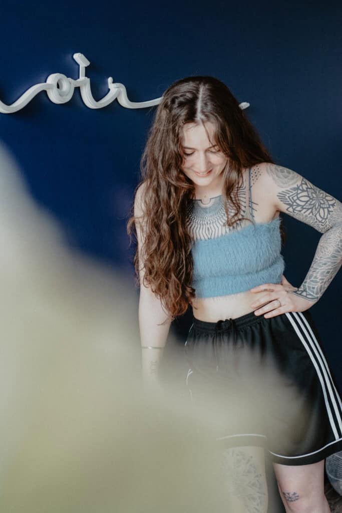 Clara vor blauer Wand mit Blackwork-Brust-Tattoo im Atelier Noir nu Köln